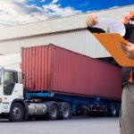 Gestão Fiscal para Transportadoras - homem conferindo documentos fiscais em frente à uma carreta vermelha e branca de 3 eixos